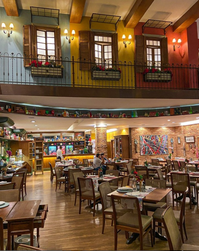 Interior de um restaurante com decoração colorida e temática rústica, destacando mesas arrumadas, janelas falsas com floreiras e luminárias.